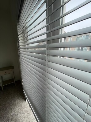 Ein Zimmer mit horizontalen Jalousien an einem Fenster, einem kleinen Holztisch und einem Teppichboden.