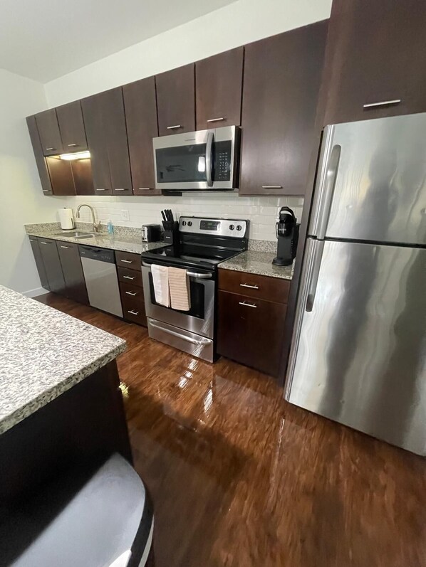 Una cocina moderna con gabinetes de madera oscura, electrodomésticos de acero inoxidable, incluyendo un microondas, estufa y refrigerador, y encimeras de granito.