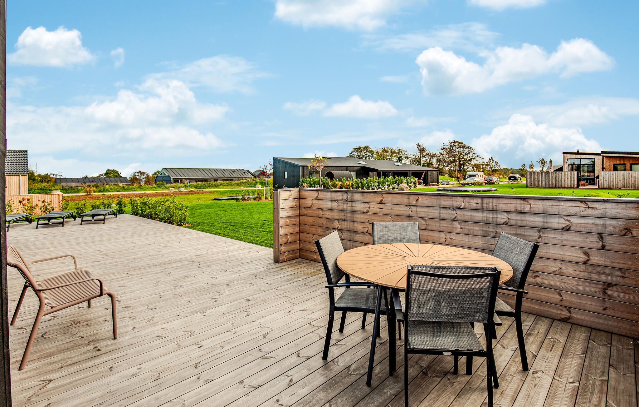 An outdoor patio with a wooden deck, a round table with chairs, and a view of a green landscape and buildings in the background.