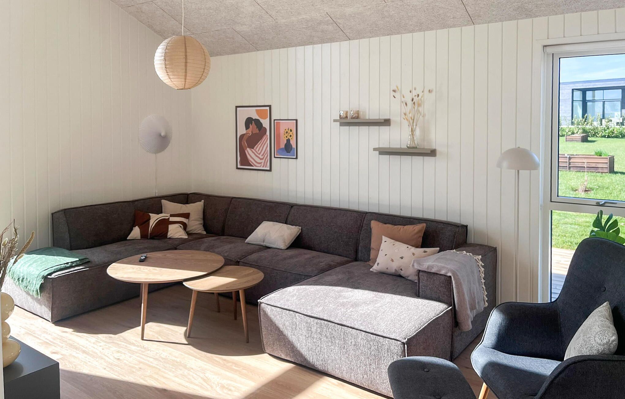 Ein modernes Wohnzimmer mit einer Eckcouch, zwei runden Couchtischen, einem Beistelltisch und einem Sessel. An der Wand sind Regale mit Dekorationen angebracht und ein großes Fenster bietet einen Blick ins Freie.