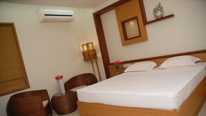 A neatly arranged bedroom with a wooden bed, a small bedside table with a vase of flowers, a chair, and a wall-mounted air conditioner. - Hotel Rithanns (Sulur)