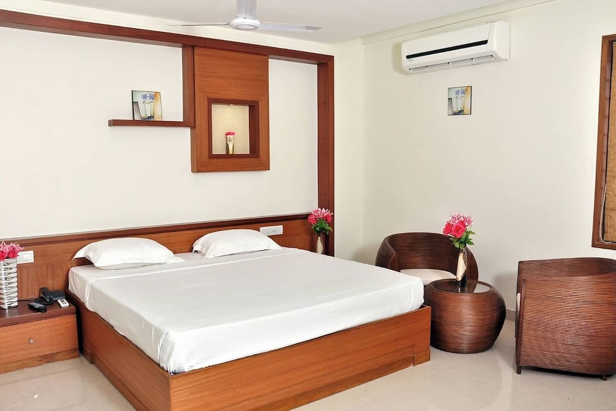A hotel room with a large bed, wooden headboard, bedside tables, a chair, a small table, and an air conditioning unit.