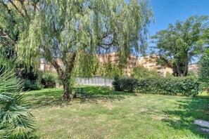 A garden with a bench, a tree, and a building in the background. - A/C 2bdr/6P flat just two minutes from the beach! (Antibes)