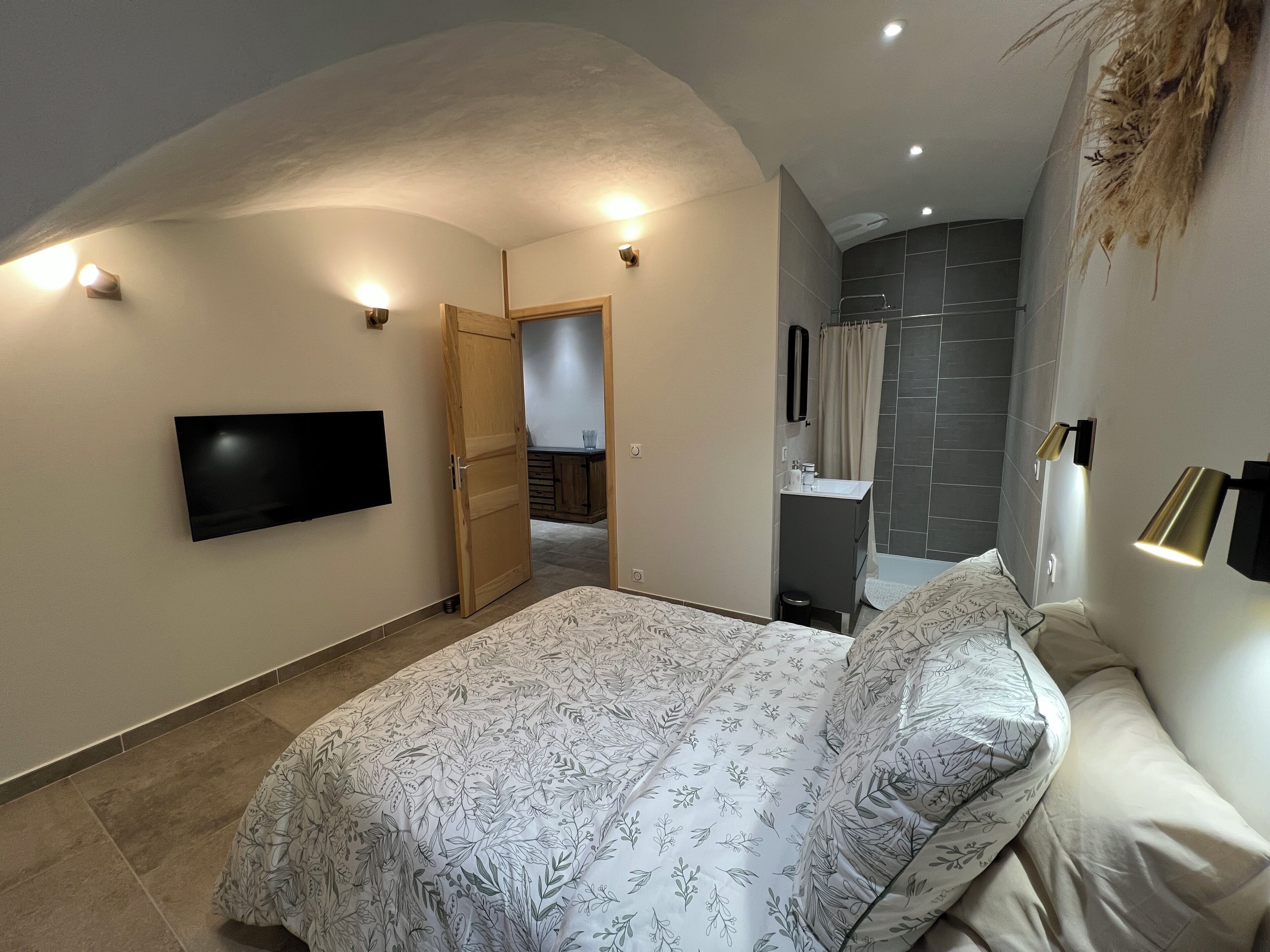 A bedroom with a bed, a TV mounted on the wall, a bathroom with a shower, and a door leading to another room.