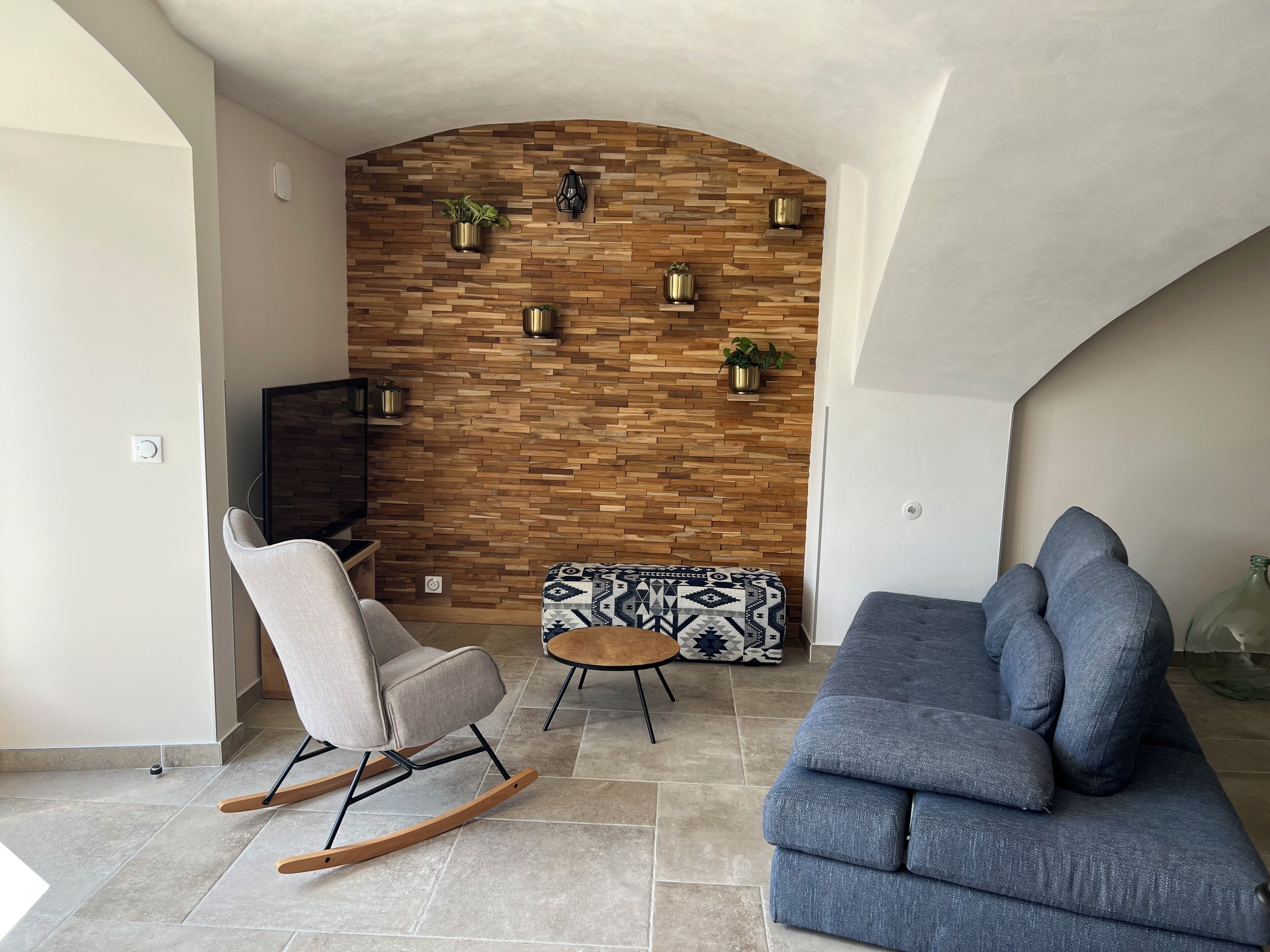 Ein Wohnzimmer mit einer Holzwand, einem grauen Sofa, einem Schaukelstuhl und einem gemusterten Hocker.