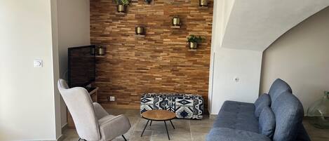 Ein Wohnzimmer mit einer Holzwand, einem grauen Sofa, einem Schaukelstuhl und einem gemusterten Hocker.