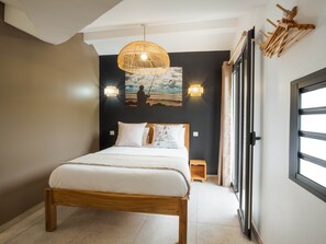A bedroom with a wooden bed, two bedside tables, a wall-mounted lamp, and a large window. - Appt Tradition - L'Escale Bleue Saint-Leu, Réunion (Saint-Leu)