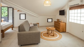 Ein modernes, helles Zimmer mit einem grauen Sofa, einem kleinen Holztisch, einem an der Wand montierten Fernseher und einem großen Fenster mit Jalousien.