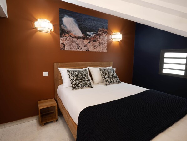 A bedroom with a wooden headboard, two wall-mounted lights, a bed with white bedding and patterned pillows, a small wooden nightstand, and a painting of a rocky coastline. - Appt Volcanik - L'Escale Bleue Saint-Leu, Reunion Island (Saint-Leu)