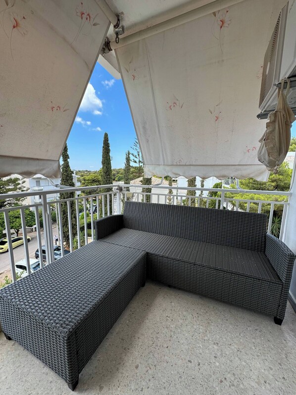 Een balkon met een rieten bank, een witte leuning en uitzicht op de straat en bomen.