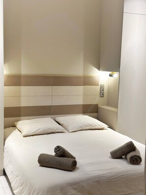 Habitación de hotel con una cama, dos almohadas, cabecero, mesita de noche y una lámpara fijada a la pared.