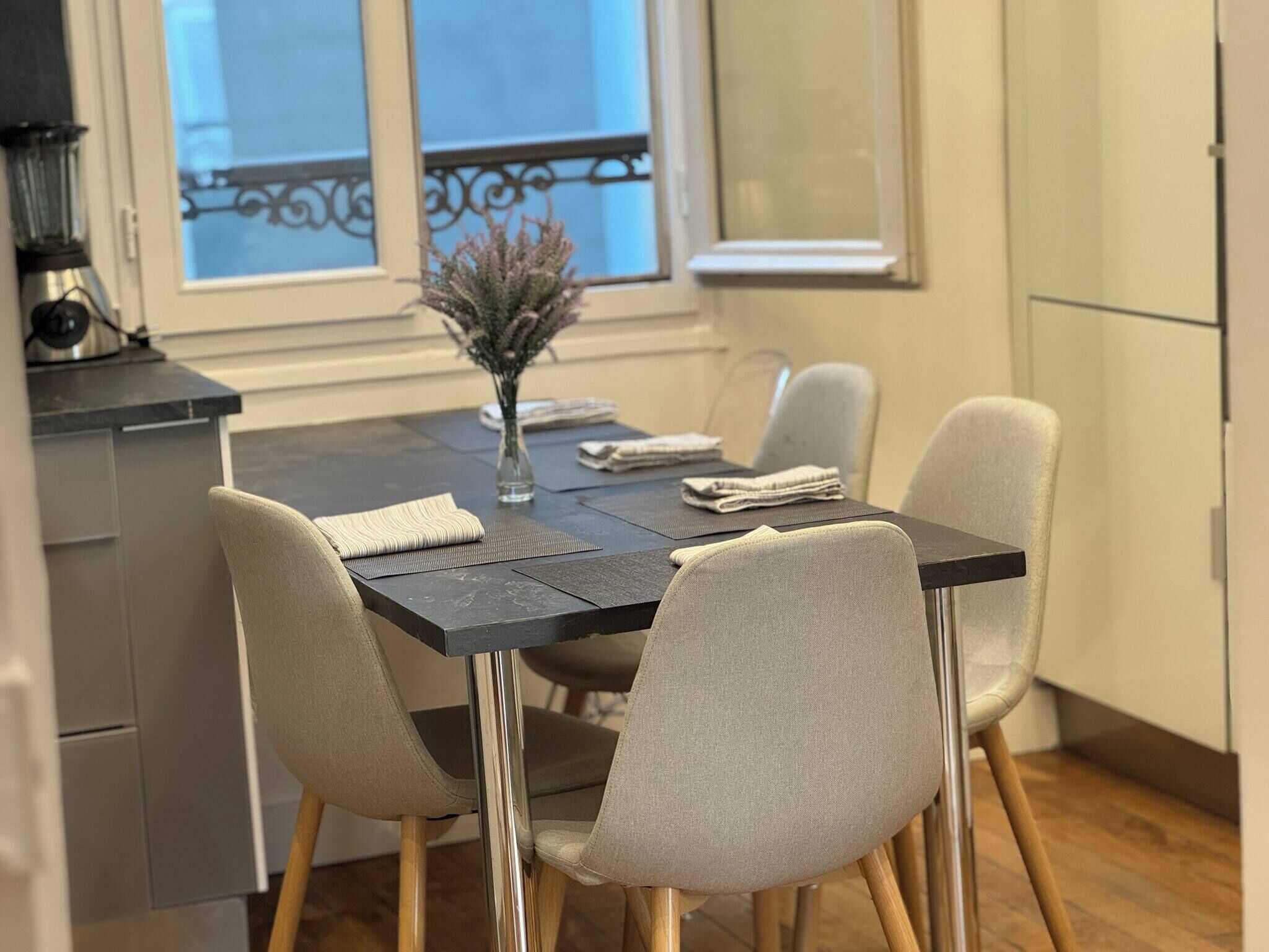 A dining area with a dark table, four chairs, a vase with flowers, and a window with a balcony view.
