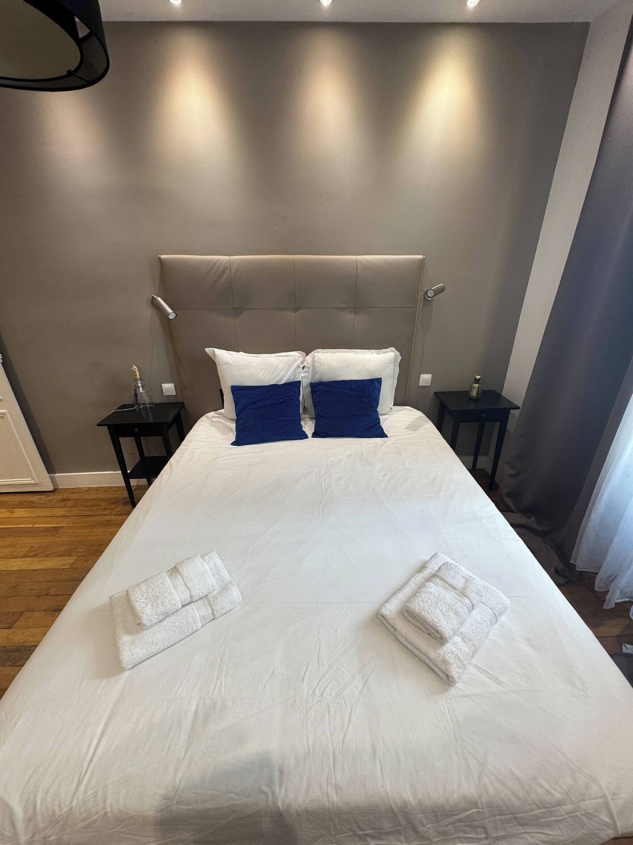 A neatly made bed with white and blue pillows, a grey upholstered headboard, and two bedside tables with lamps.