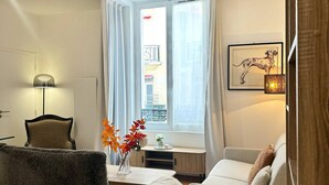 Een woonkamer met een bank, fauteuil en een tafel met een bloemenvaas. Er is een raam met gordijnen, een lijst met een foto aan de muur en een lamp op een bijzettafeltje.