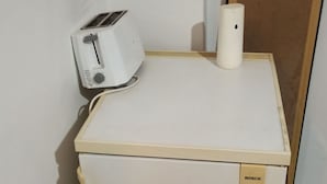 Un petit réfrigérateur blanc de la marque Bosch, surmonté d’un grille-pain, est placé dans un coin d’une pièce dont le sol est recouvert de carreaux.