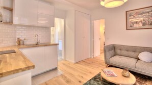 Cocina moderna con armarios blancos, encimera de madera y lavaplatos integrado. En la zona de estar, hay un sofá y una mesa de centro, y un cuadro en la pared.