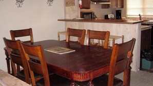 Een eetgedeelte met een houten tafel en stoelen, een keuken op de achtergrond en een brandblusser aan de muur.