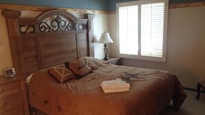 Een slaapkamer met een houten hoofdbord, een bed met bruin beddengoed, een nachtkastje met een lamp en een raam met jaloezieën.