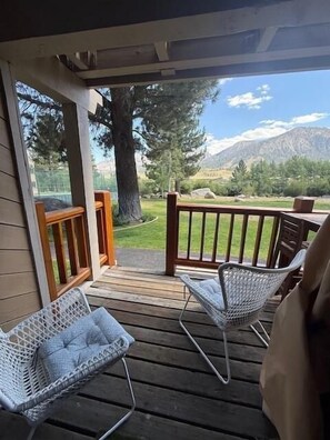 Une terrasse extérieure couverte, dotée de deux fauteuils en osier et d’une table en bois, qui offre une vue sur une zone herbeuse, des arbres et des montagnes en arrière-plan.
