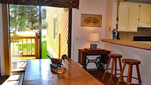 A wooden dining table with a bird figurine, a lamp, and two stools in a kitchen area with a view of a garden through a sliding door. - Mammoth Creek Unit 45 (Mammoth Lakes)