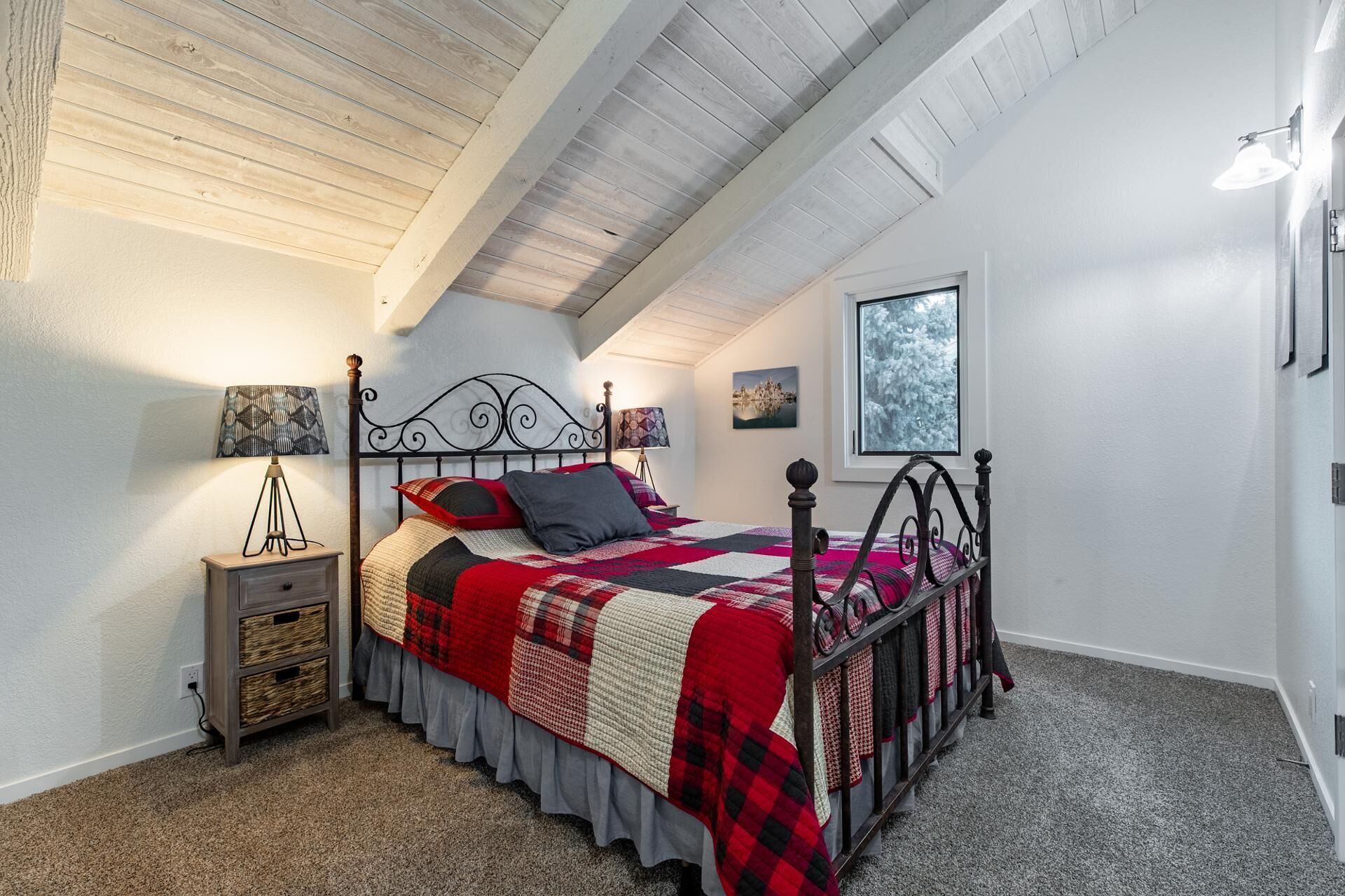 Ein Schlafzimmer mit geneigter Decke, einem Bett mit rot-schwarzer Steudecke, zwei Nachttischlampen, einem Nachttisch mit Aufbewahrungskörben und einem Fenster mit Blick auf Bäume.