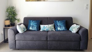 Ein graues Sofa mit vier gemusterten Kissen, ein an der Wand hängendes Bild von einem Strand, eine Bambuspflanze auf einem Beistelltisch und ein gefliester Boden.