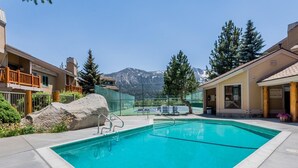 Ein Schwimmbecken mit Bergblick, umgeben von Gebäuden mit Balkonen und einem Tennisplatz.