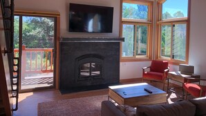 A living room with a fireplace, a television mounted above it, and large windows offering views of the outdoors. - Mammoth Creek Unit 12 (Mammoth Lakes)