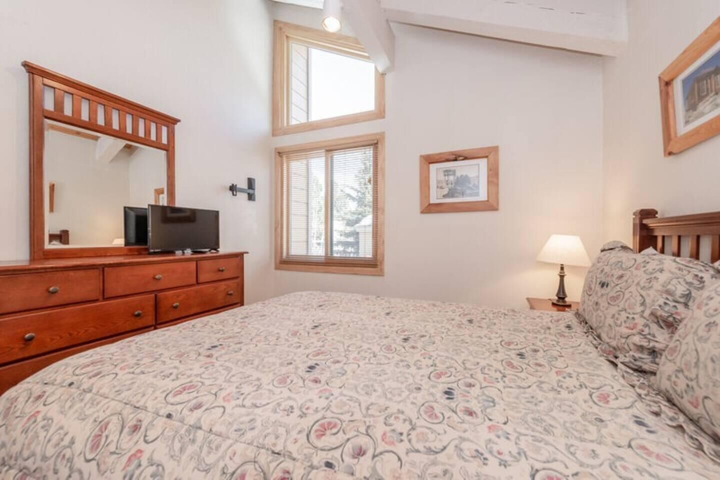 Ein Schlafzimmer mit einem großen Bett, einer Holzkommode, einem Fernseher, einem Fenster mit Jalousien und einem gerahmten Bild an der Wand.
