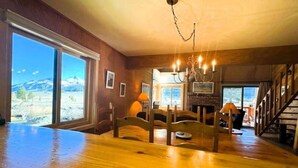 A spacious room with a wooden floor, large windows offering a mountain view, a fireplace, and a staircase leading to another level. - Mammoth Creek Unit 32 (Mammoth Lakes)