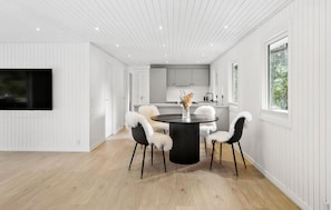 A modern interior with a dining area featuring a round table and chairs, a kitchen with white cabinets, and a large window providing a view of greenery.