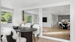 A modern interior with a dining area featuring a round table and chairs with white fur throws, a kitchen with a flat-screen TV, and a large sliding glass door leading to a balcony. - Nice home in Farsø with house a panoramic view (Farsø)