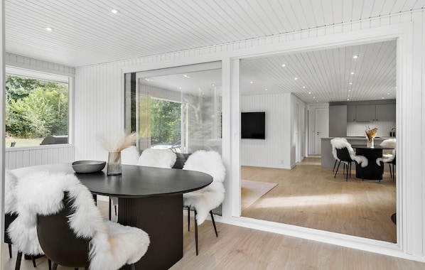 A modern interior with a dining area featuring a round table and chairs with white fur throws, a kitchen with a flat-screen TV, and a large sliding glass door leading to a balcony.