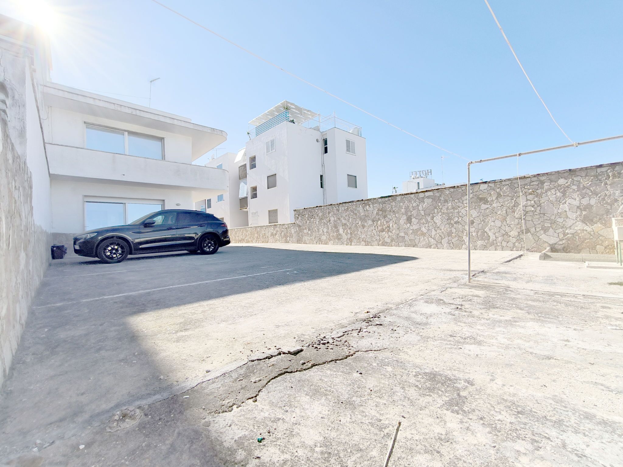 Un aparcamiento con un coche negro, un edificio blanco con balcones y una pared de piedra.