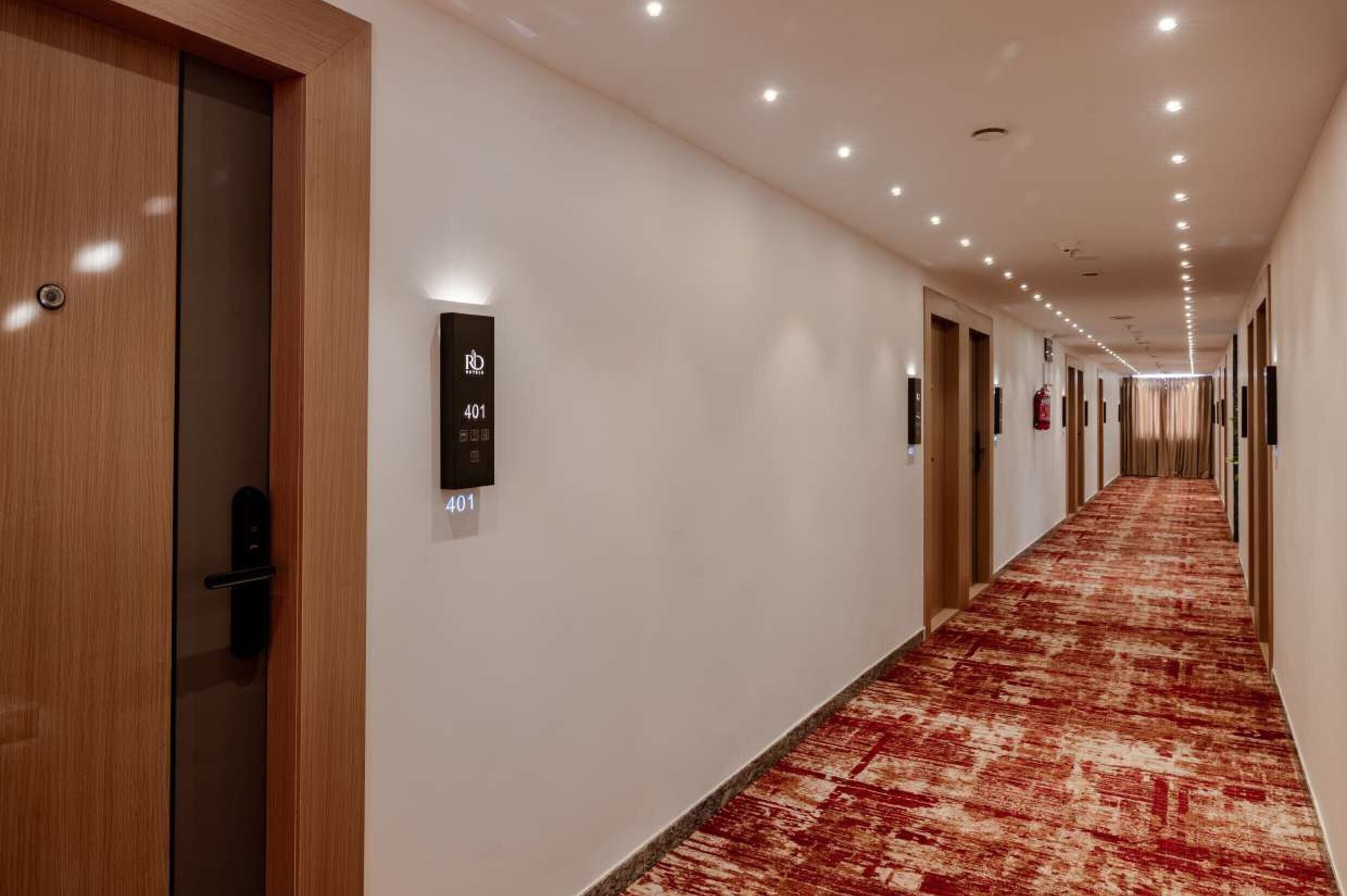 Un pasillo de hotel con alfombra roja, puertas de madera y letreros con números que señalan la ubicación de las habitaciones.