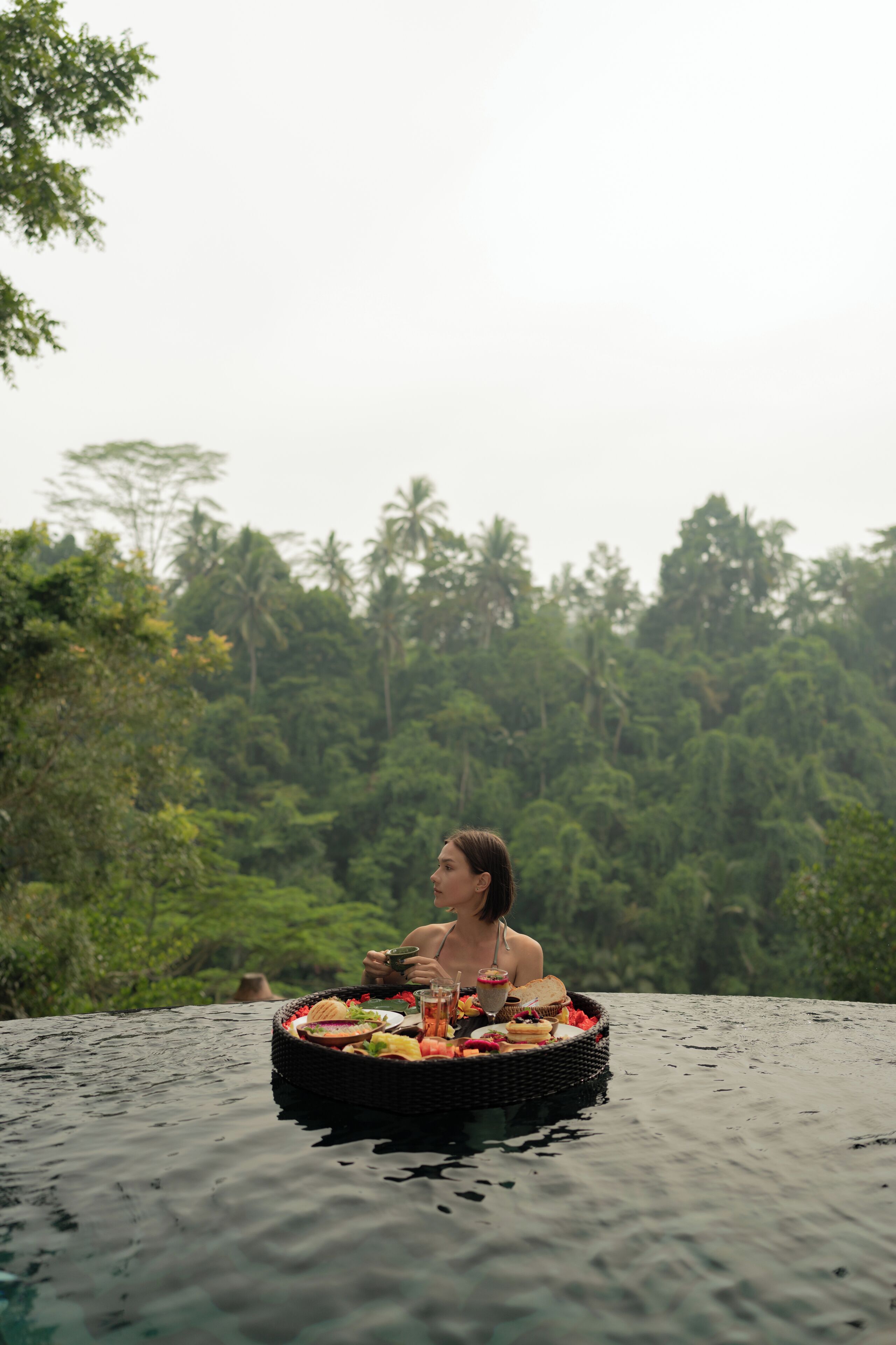A person relaxing on a floating platform with a basket of food and drinks, surrounded by lush greenery.