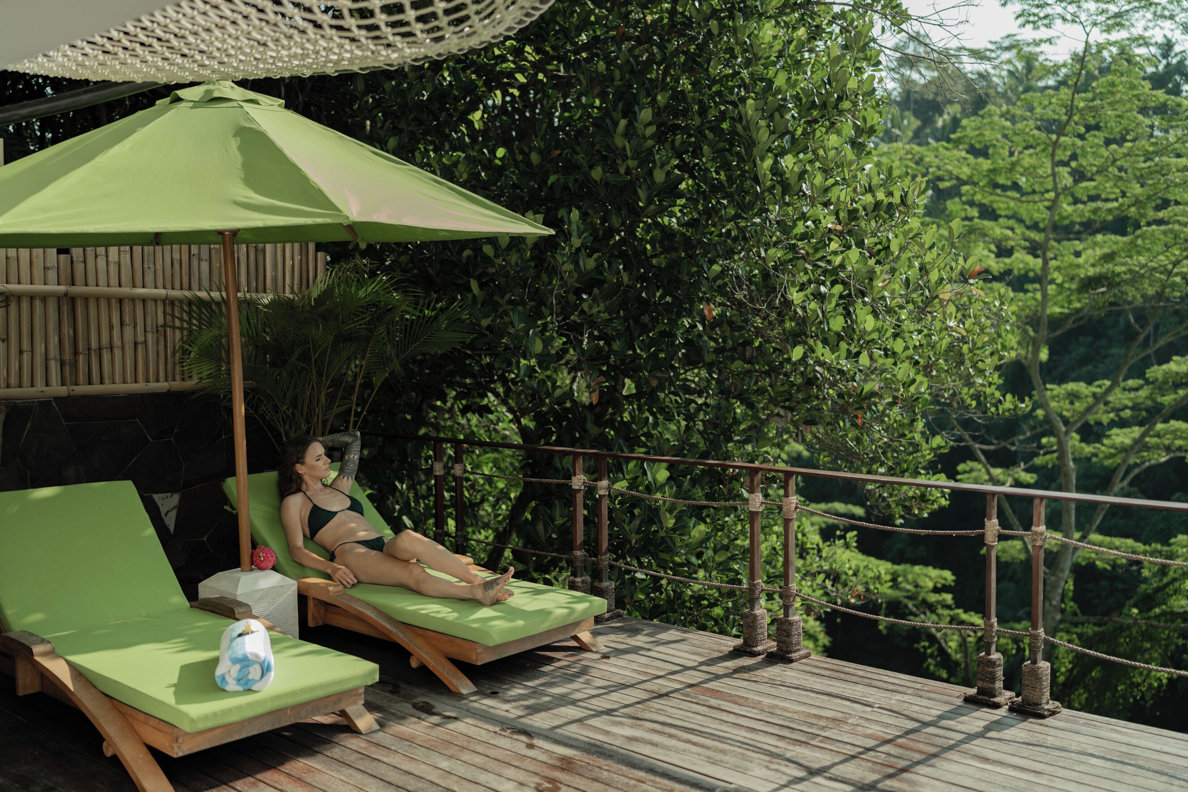 Uma pessoa relaxando em um deck de madeira com um guarda-sol verde, rodeada por vegetação exuberante.