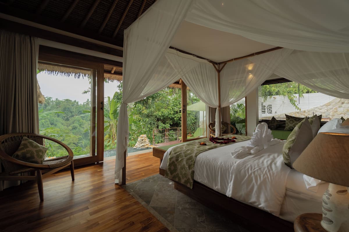 A bedroom with a canopy bed, wooden flooring, and a view of lush greenery outside.