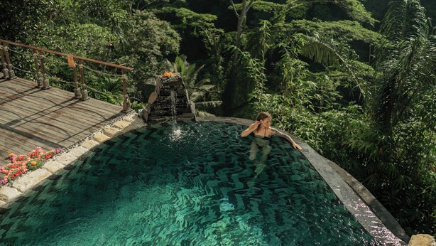 A pool with a wooden deck and a waterfall feature, surrounded by lush greenery. - Marangga ubud valley view & private pool (Ubud)
