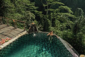 A pool with a wooden deck and a waterfall feature, surrounded by lush greenery. - Marangga ubud valley view & private pool (Ubud)