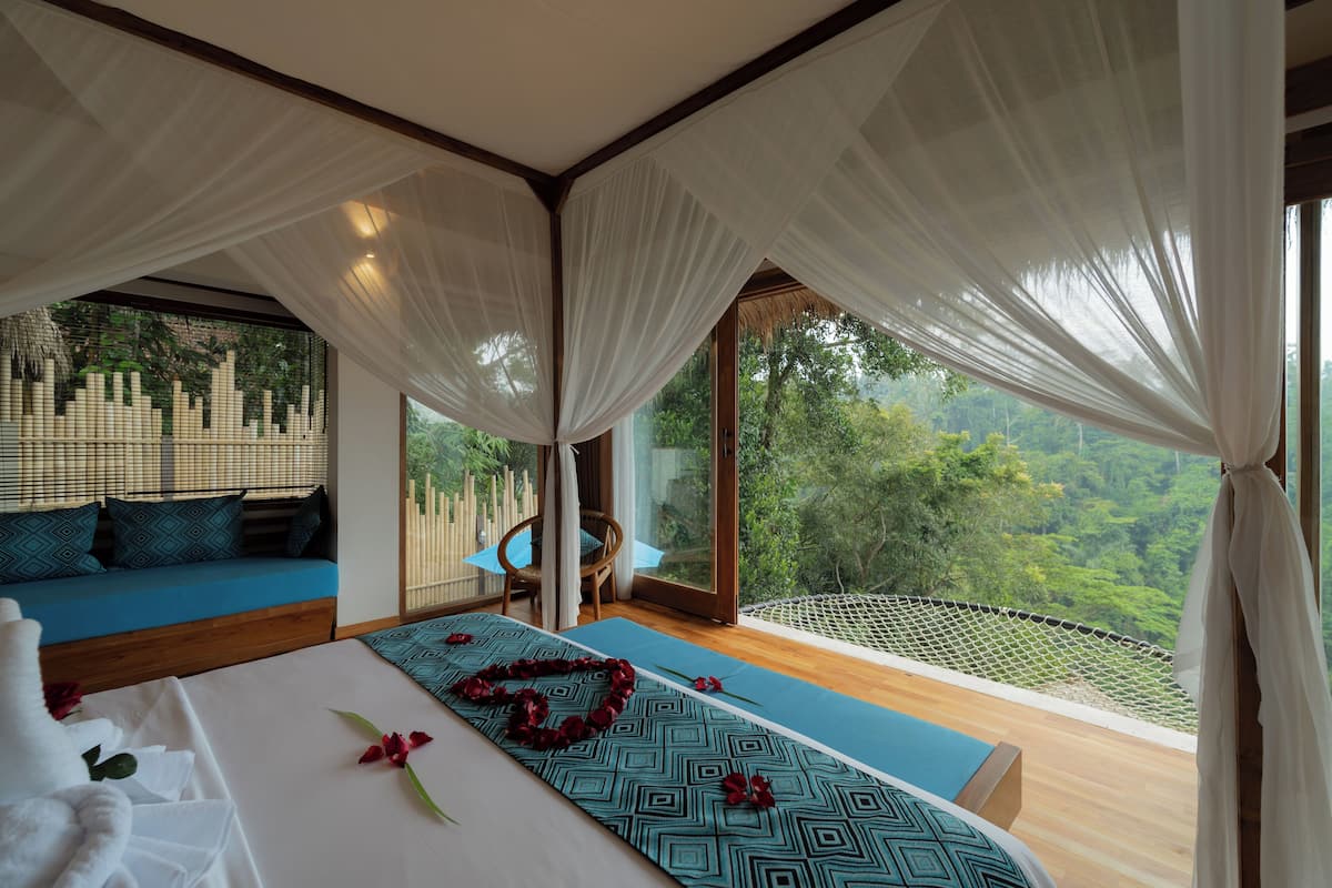 A bedroom with a large bed, a blue patterned sofa, and a view of lush greenery.
