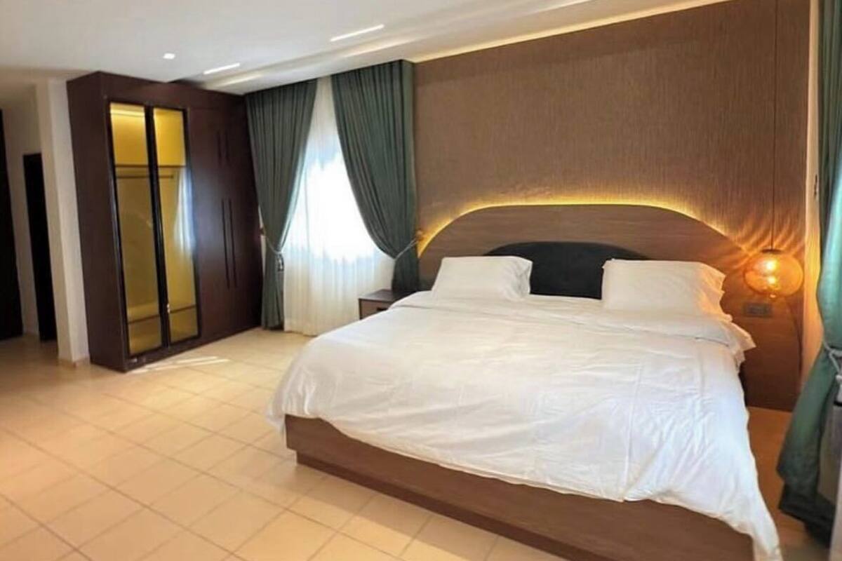 A large bed with a wooden headboard, a bedside lamp, and a view of a room with a wardrobe and curtains.