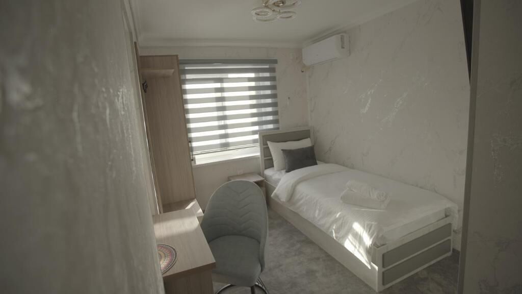 A small, single-bed room with a desk and chair, a window with blinds, and a ceiling fan.