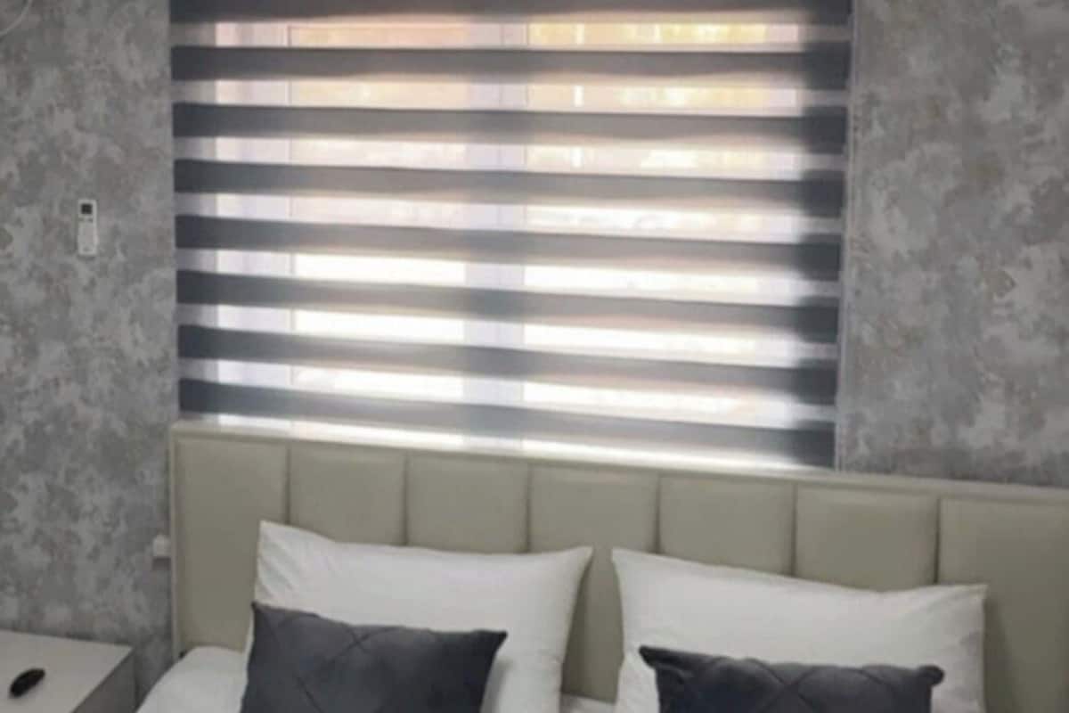 A bed with white bedding and two dark pillows, a bedside table with a phone, and a window with blinds.