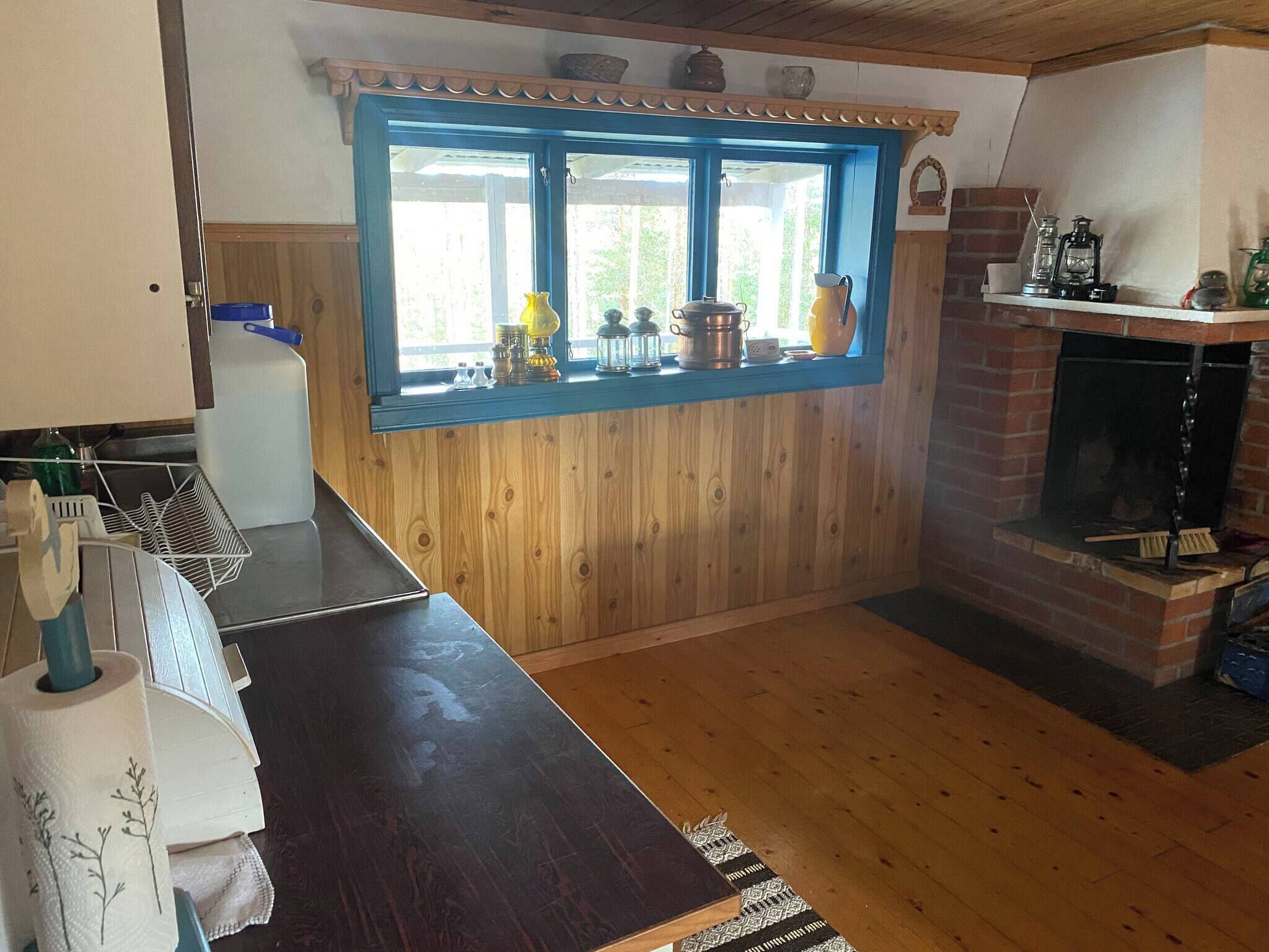 A rustic kitchen with wooden walls, a blue-framed window, and a brick fireplace.