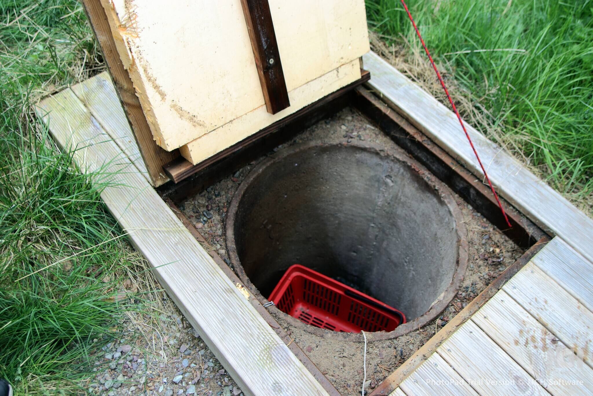 A manhole with a red plastic basket inside, surrounded by wooden planks and grass.