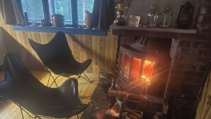 A cozy room with a wood-burning stove, two black chairs, a wooden floor, and a window overlooking a forest. - Off Grid at Fjätälven (Sarna)