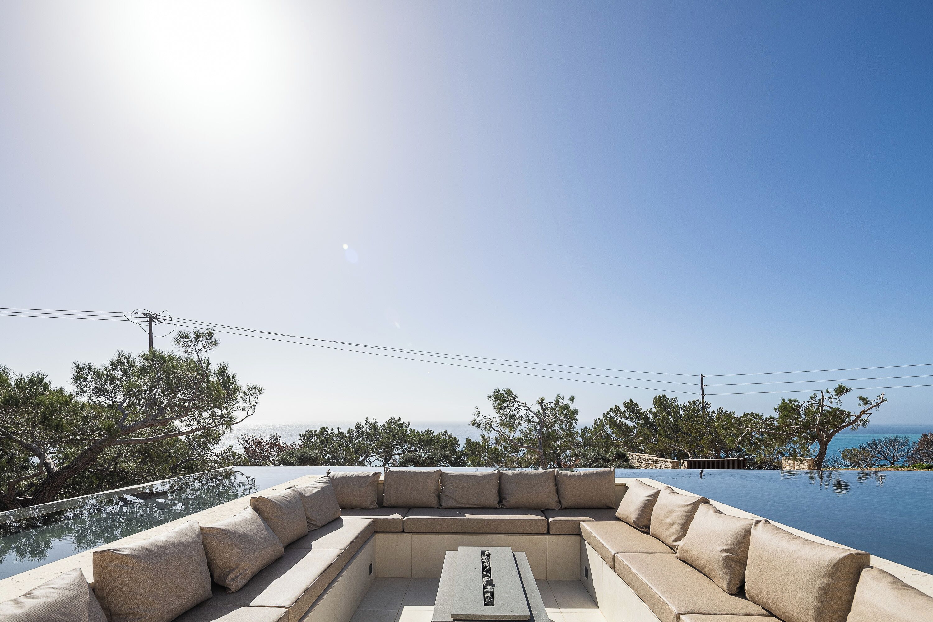 Piscina infinita con sofá seccional color beige y una mesa, con vista a un paisaje costero con árboles y el mar.