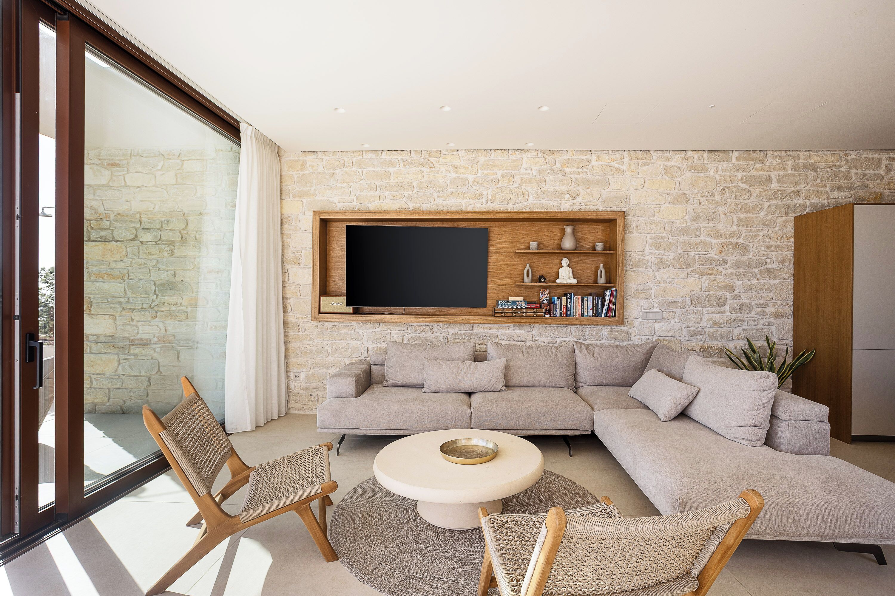 A modern living room with a stone wall, a large flat-screen TV, a beige sofa, and wicker chairs.