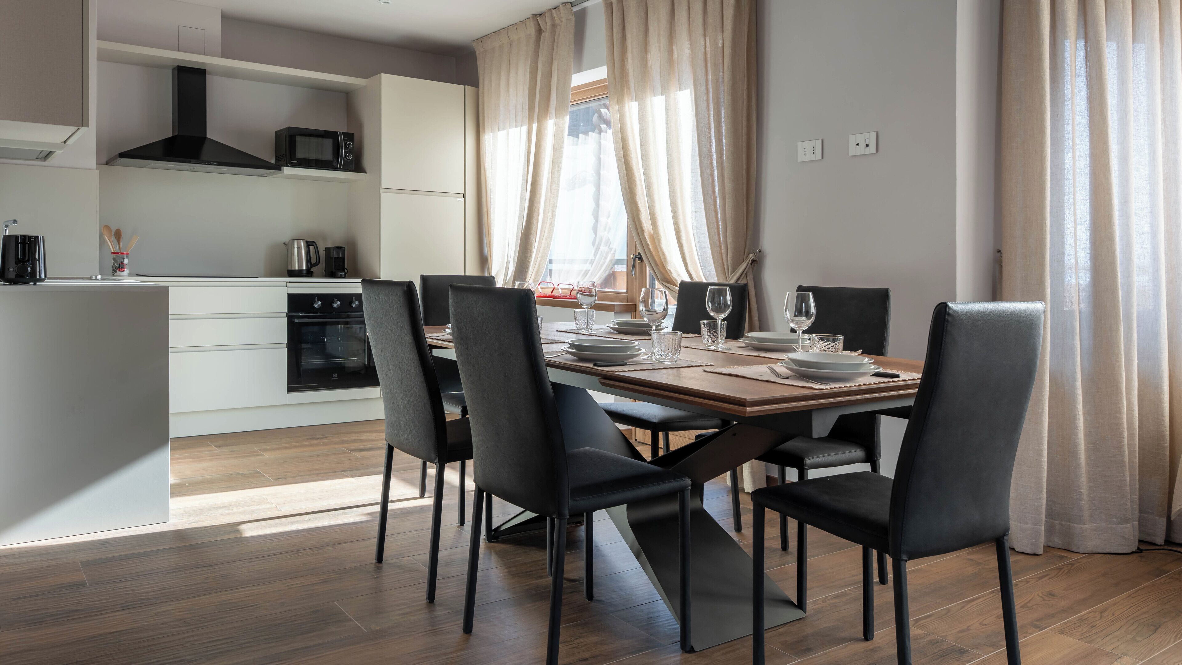 Een moderne keuken met witte kasten, een zwarte magnetron en een kookplaat. Er is een eethoek met een houten tafel gedekt voor vier personen, omringd door zwarte stoelen.